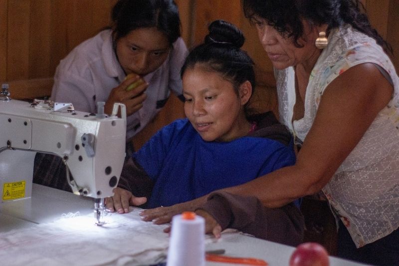 Three women work together on an electric sewing machine, sewing a white fabric