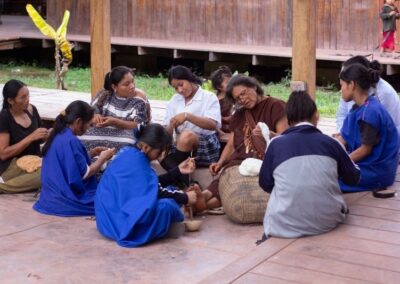 A group of indigenous women and girls sits in a circle on a wooden veranda, spinning or weaving wool