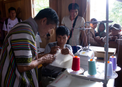Two men are working at a sewing mechine with colorful spools of yarn, while other people in the background watch the process attentively